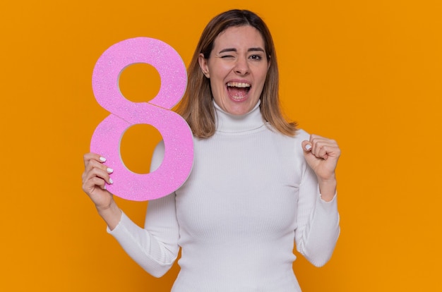happy and excited young woman in white turtleneck holding number eight made from cardboard