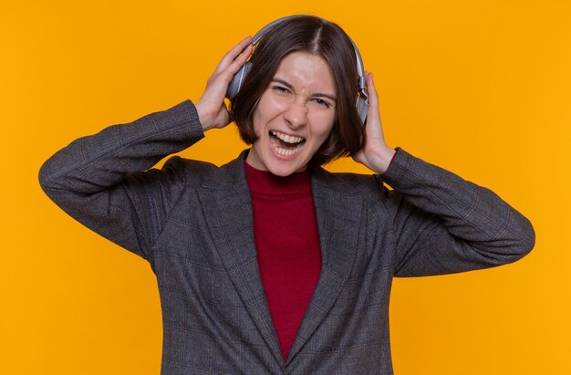 Happy and excited young woman with short hair wearing grey jacket with headphones enjoying her favorite music smiling cheerfully standing over orange wall