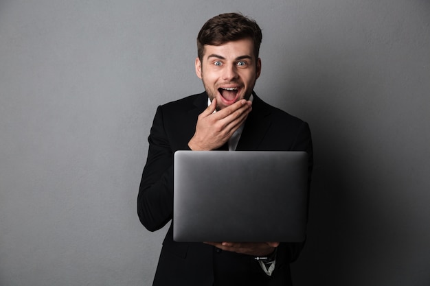 Happy exited businessman covering his mouth while holding laptop