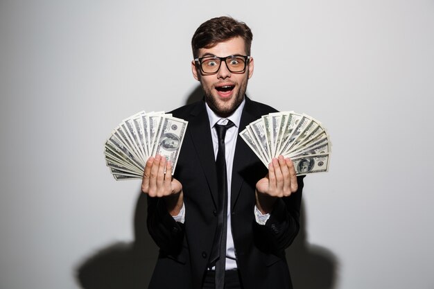 Счастливый возбужденный мужчина в очках и черном костюме держит две кучки денег