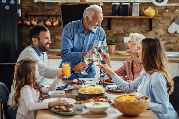 Счастливая большая семья наслаждается обедом и тостами за обеденным столом. В центре внимания пожилая пара