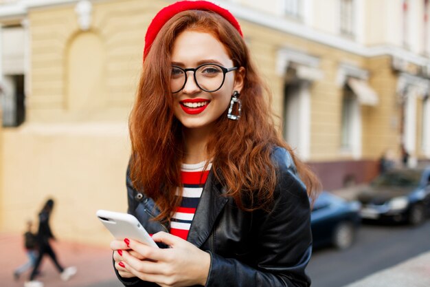 Счастливая сказочная рыжая женщина в стильном красном берете на улице с помощью смартфона