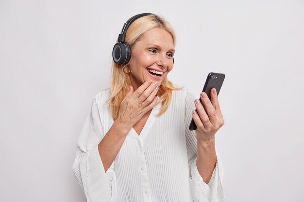 Счастливая белокурая взрослая женщина делает онлайн-звонок, используя смартфон и наушники, рада слышать, что лучший друг изучает новые технологии, смотрит видео на мобильном устройстве, изолированном на белой стене студии
