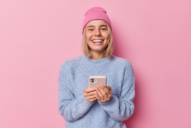 幸せな金髪の女性技術ユーザーは、ピンクのスタジオの背景に対して帽子とジャンパーのポーズを身に着けているボーイフレンドとオンラインでチャットすることを喜んでスマートフォンを保持しますセルラー技術とネットワークの概念