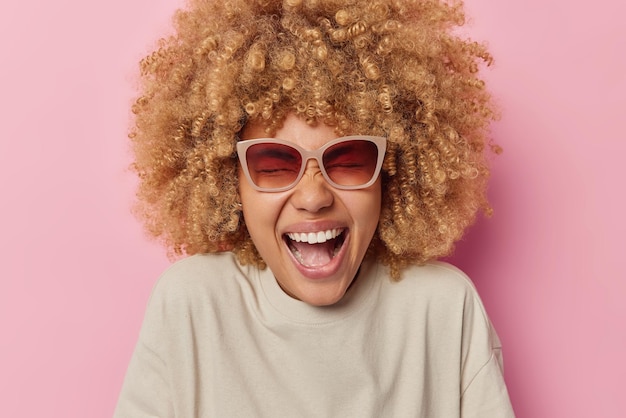 Free photo happy fair haired teenage girl laughs joyfully chuckles at camera stands carefree keeps mouth opened wears sunglasses casual clothes isolated over pink background positive emotions concept