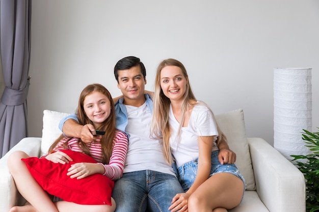 Счастливая семья на диване в гостиной, включая отца, мать и дочь, наслаждаются просмотром телевизора вместе с пультом дистанционного управления