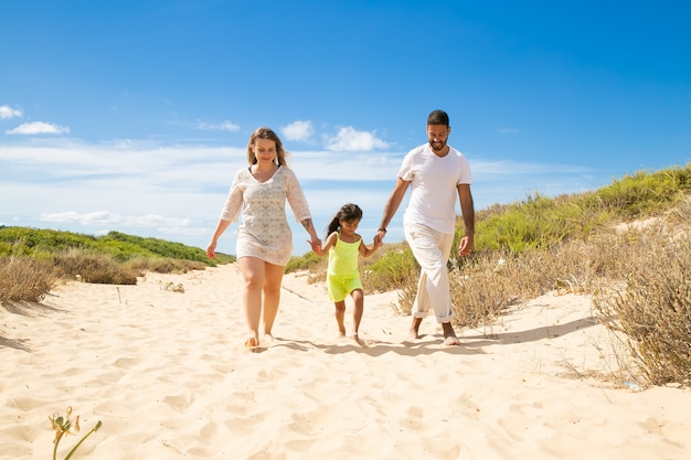 Happy family couple and little kid in summer clothes walking white along sand path, girl holding parents hands