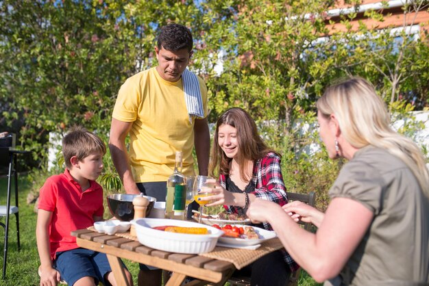 裏庭でバーベキューを食べる幸せな家族。中年の大人の親と子供がテーブルの周りに座って、作りたての食べ物を食べ、飲み物を飲みます。バーベキュー、料理、食べ物、家族のコンセプト