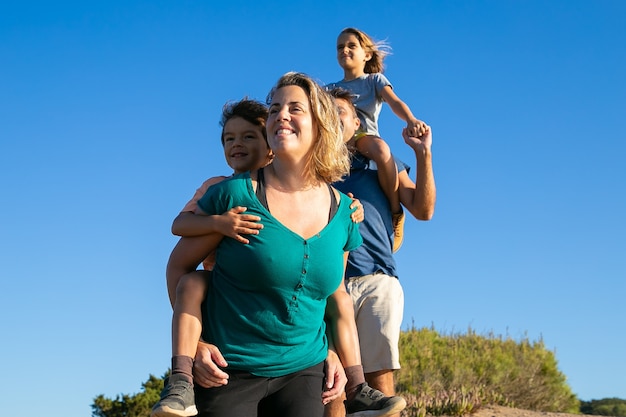 Happy family enjoying hiking in countryside. Two kids riding on parents back and neck. Low angle. Nature and recreation concept