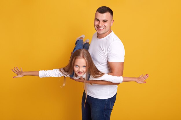 happy family father and daughter, child desses denim onalls pretending being plane with their hands spreading aside and having fun with her daddy, isolated on yellow wall