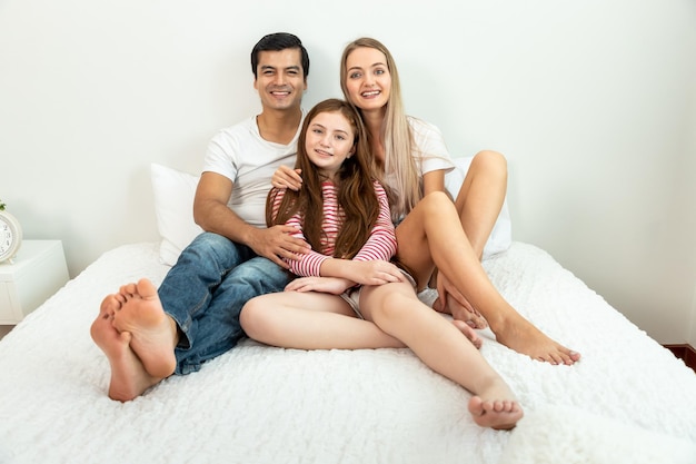 Happy family including father mother and daughter are sitting on bed with smile looking at camera