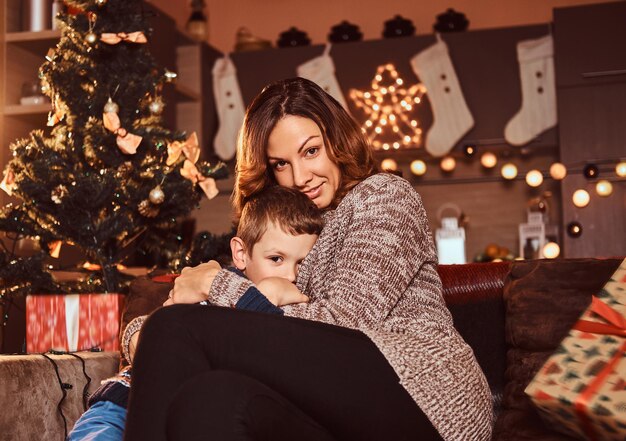 Happy family. Mom hugging her cute little boy while sitting on sofa in decorated room during Christmas time.