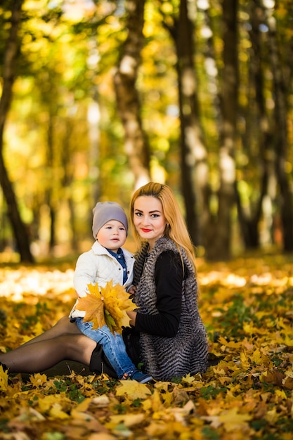 黄色の葉の上に横たわる木の近くの秋の公園で子供と遊んで幸せな家族の母。秋のコンセプトです。