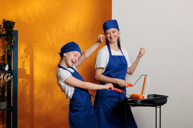 Happy family painting walls with orange color paint and roller brush, having fun with house renovation. Mother and little child using paintbrush and redecoration tools to do housework improvement.