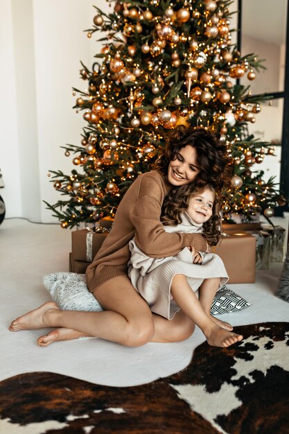 딸과 함께 예쁜 엄마의 행복한 가족 초상화는 크리스마스 트리 앞에 앉아 새해를 축하하는 니트 스웨터를 입고