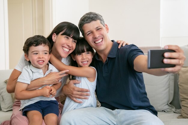 Happy family with two little kids sitting on couch at home together, taking selfie