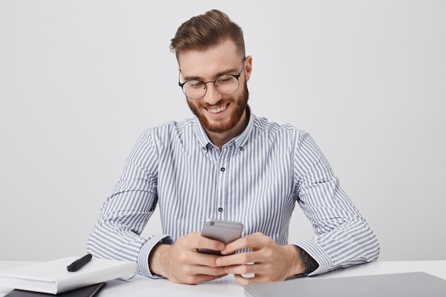 Happy fashionable male student sits on work place, prepares for classes, holds mobile phone