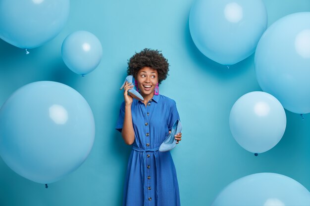 幸せなファッショナブルな女性は誕生日の休日のパーティーの準備をし、電話として耳の近くにハイヒールの靴を持ち、お祝いの服を着て、楽しんで、ポーズをとる