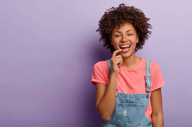Happy fashionable woman smiles carefree, wears t shirt and denim overalls, keeps fore finger on lips, isolated over purple background