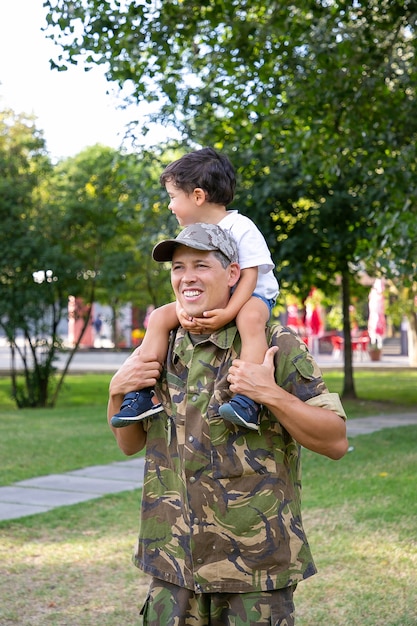 Happy father holding son on neck and holding his legs. Cheerful Caucasian dad in uniform walking with little boy in park. Cute boy looking away. Family, fatherhood and returning home concept