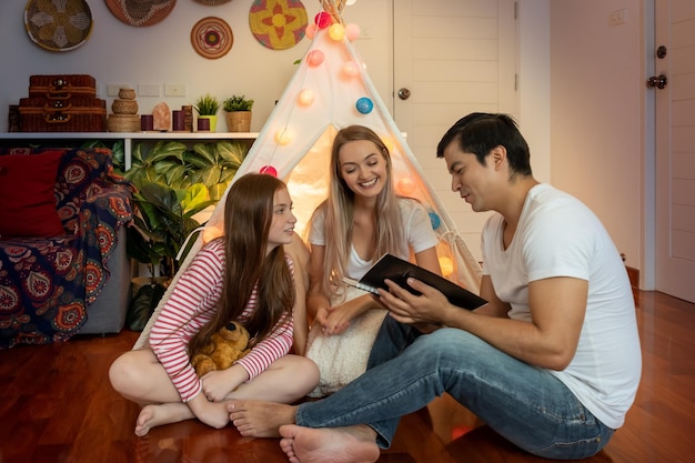 Happy father and mother reading book and telling story with daughter in front of tepee tent in bedroom family relationship concept