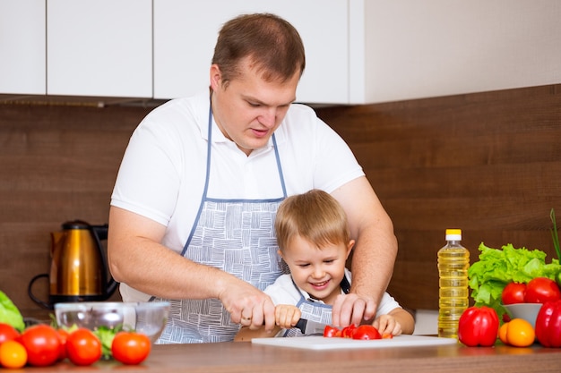 A happy father and a young son prepare a salad in the kitchen with vegetables. My dad teaches me how to cut tomatoes on a blackboard. Concept of diet food