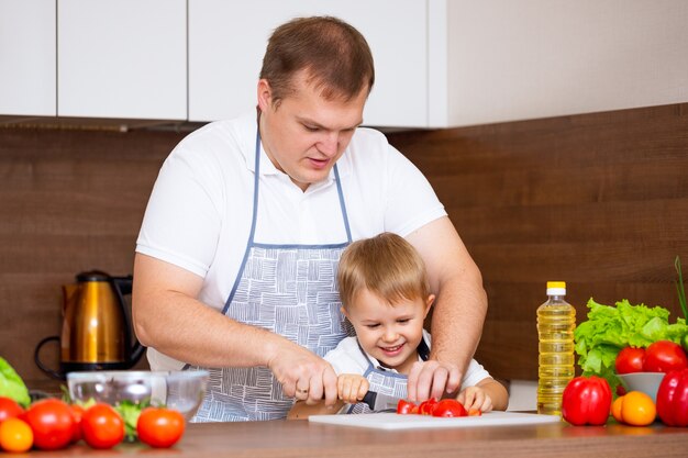 A happy father and a young son prepare a salad in the kitchen with vegetables. My dad teaches me how to cut tomatoes on a blackboard. Concept of diet food