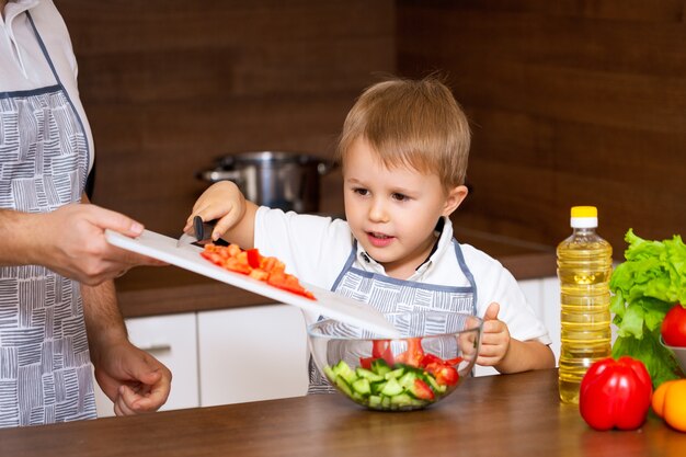 幸せな父と幼い息子が野菜を使ってキッチンでサラダを準備します。私の父は黒板にトマトを切る方法を教えてくれます。ダイエット食品のコンセプト