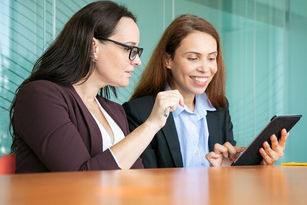 Happy female business colleagues using tablet together, looking at screen and smiling while sitting at table in meeting room.