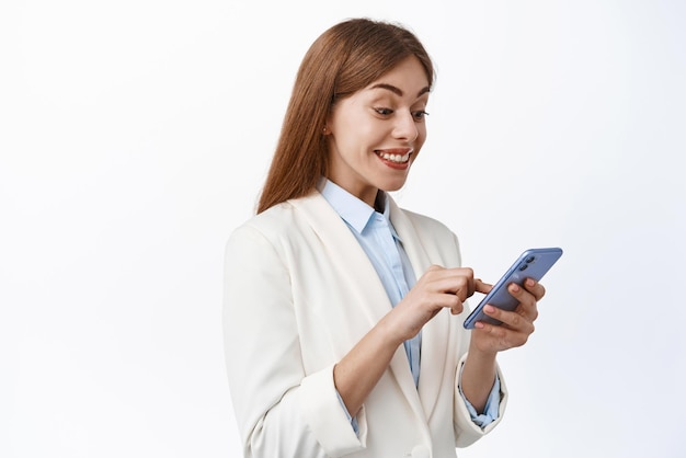 Happy female ceo manager in business suit looks at smartphone screen and smile pleased reading message on phone standing over white background
