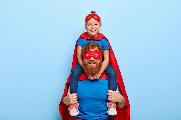 Free photo happy female child sits on fathers shoulders, feels heroic and strong, wears red mask