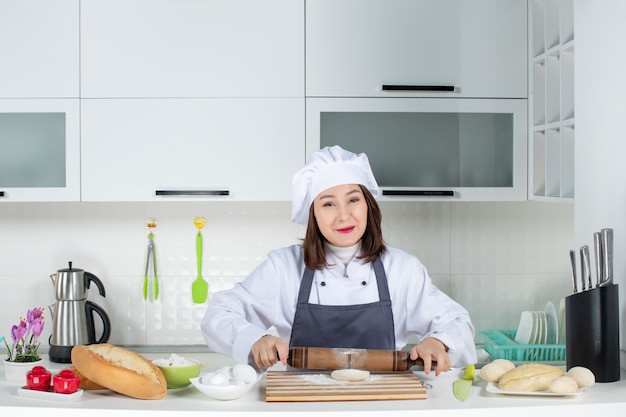 Felice chef commis femminile in uniforme in piedi dietro il tavolo che prepara la pasticceria nella cucina bianca