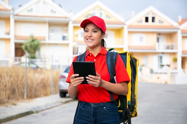 Happy female courier delivering order and working in post service. Smiling deliverywoman in red cap and shirt carrying yellow backpack and holding tablet. Delivery service and online shopping concept