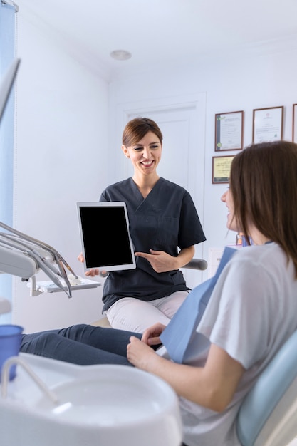 Happy female dentist pointing on digital tablet screen to female patient in clinic