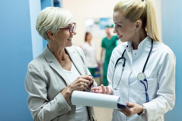 Happy female doctor and mature woman talking while analyzing medical reports in the hospital