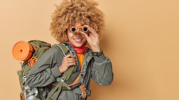 Free photo happy female explorer observes something far away uses binoculars observes spectacular view discovers new places carries rucksack isolated over brown background blank space for your promotion