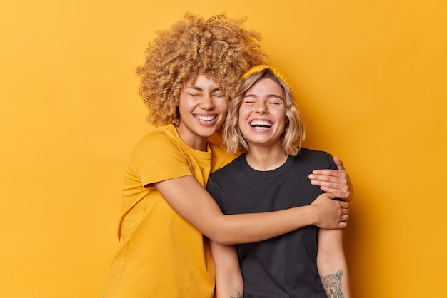 Happy female friends embrace and have good relationshipes laugh positively dressed in casual t shirts keep eyes closed isolated over yellow background glad sisters love each other stand indoor