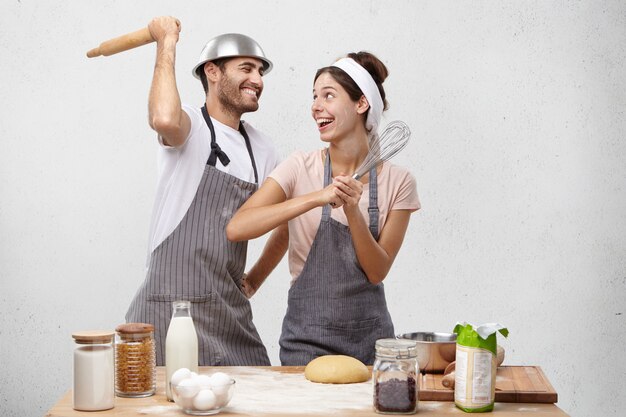 Happy female and male foolish at kitchen, fight with whisk and rolling pin, have glad expressions