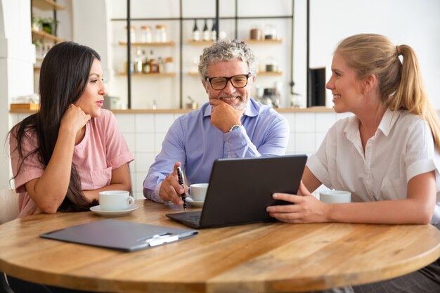 Happy female manager presenting project on laptop to young woman and mature man, discussing content with satisfied customers