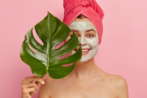 幸せな女性モデルは、フェイシャルマスクでディープクレンジングを行い、顔の半分を緑の葉で覆い、彼女の外観を改善し、素晴らしい肌を持ちたい、毛穴の詰まりを取り除き、優しく微笑む