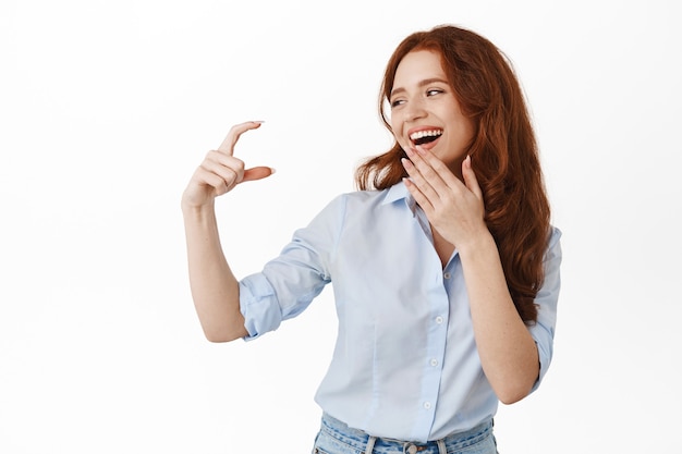 Felice modello femminile che guarda una piccola cosa, mostrando un piccolo gesto della mano e ridendo, sorridendo compiaciuto, in piedi in camicetta su bianco