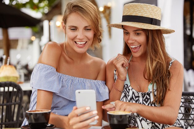 Happy female models rest together in cafeteria, surf social networks on cell phone, drink coffee or cappuccino. Joyful brunette young woman indicates at mobile phone, shares photos with friend