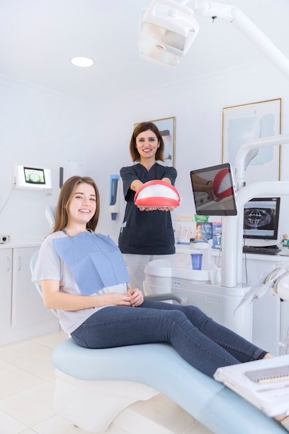 Happy female sitting on dental chair in front of dentist holding teeth model