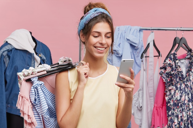 Happy female standing at clothing store, messaging with friend over smart phone while trying new clothes asking for advice what to buy. Cheerful woman using modern cell phone in shopping mall.
