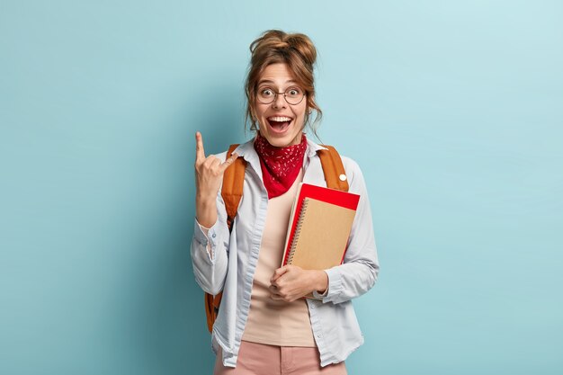 Happy female student with combed hair, makes rock n roll gesture, has glad expression, rejoices finishing of studying year