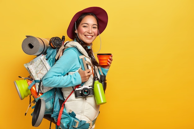 Happy female tourist drinks coffee or tea, poses with backpack, rolled sleeping rag, wears hat, jumper and vest, stops during journey, isolated over yellow wall