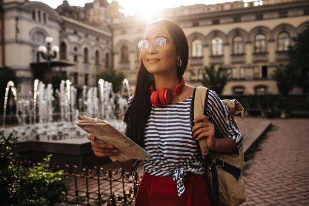 眼鏡と赤いヘッドフォンで幸せな女性の観光客は目をそらします笑顔は地図とベージュのバックパックを外に保持します