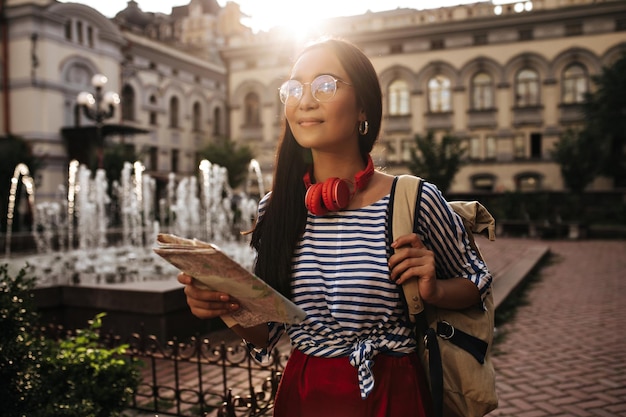 Happy female tourist in eyeglasses and red headphones looks away smiles holds map and beige backpack outside