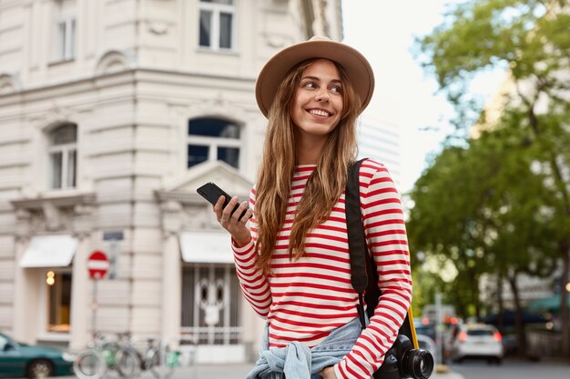 Happy female traveler carries camera for making photos, holds smart phone, texts messages online