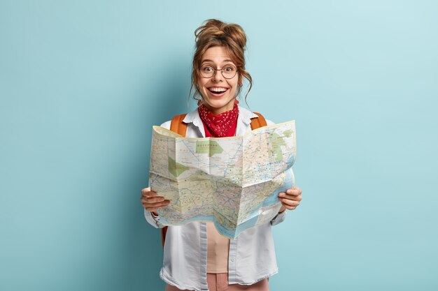幸せな女性旅行者は地図を持って、目的地のルートをチェックし、休暇中にヨーロッパを旅行し、バックパックを運び、丸い眼鏡をかけます